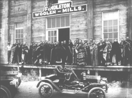 A historic black-and-white photo of a group of people in traditional attire outside Pendleton Woolen Mills, with an early car.