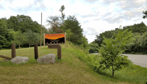 Sign for "Sunset Place" on a grassy area near a road, surrounded by trees and rocks under a cloudy sky.