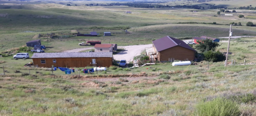 A rural landscape featuring a cluster of wooden buildings, vehicles, and laundry hanging outside on a sunny day.