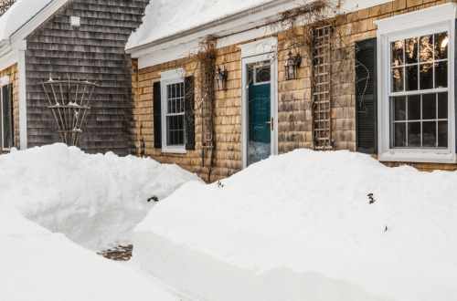 A house with wooden siding surrounded by deep snow, featuring a blue door and snow-covered pathways.