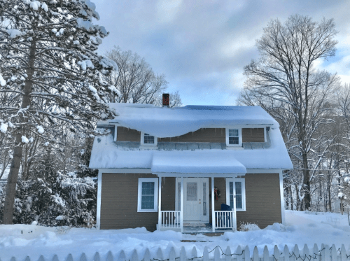 A cozy two-story house covered in snow, with a white picket fence and trees in a winter landscape.