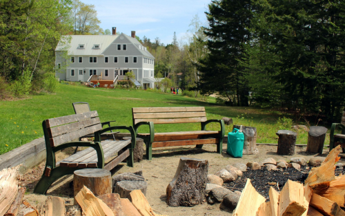 A fire pit area with wooden benches and logs, set against a grassy field and a large gray house in the background.