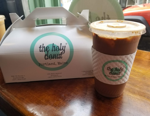 A takeout box from The Holy Donut in Portland, Maine, next to a cup of iced coffee on a wooden table.