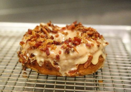 A glazed donut topped with crispy bacon bits, resting on a wire rack.