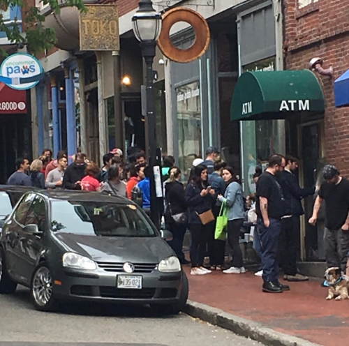 A long line of people waiting outside an ATM, with a parked car in the foreground and shops in the background.
