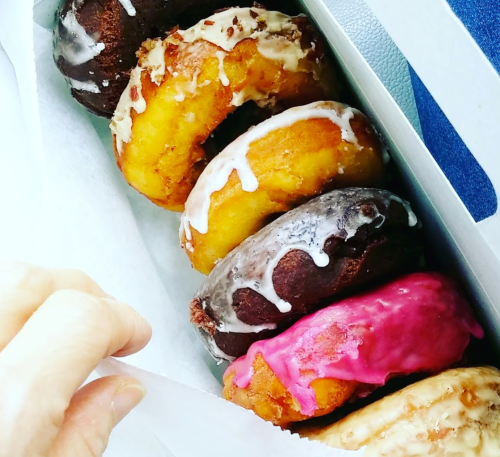 A hand reaching for a box of assorted donuts, featuring chocolate, glazed, and pink frosted varieties.