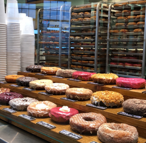 A display of colorful donuts on a wooden shelf, with various toppings and flavors, behind stacks of paper cups.