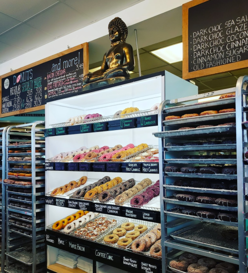 A display of colorful donuts in a shop, with a Buddha statue and chalkboard menu above.