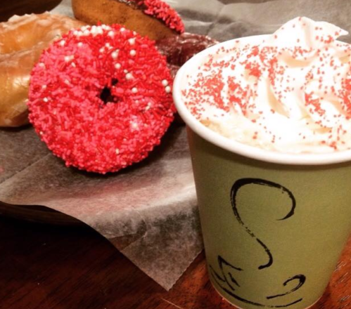 A green cup of whipped cream topped with red sprinkles, alongside colorful donuts on a wooden table.