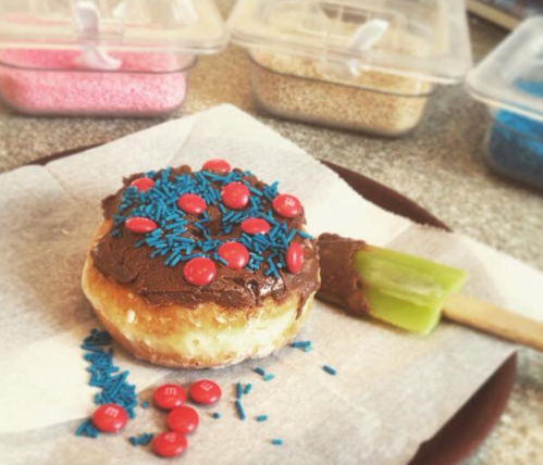 A frosted donut topped with blue sprinkles and red candies, with a green spatula beside it on parchment paper.