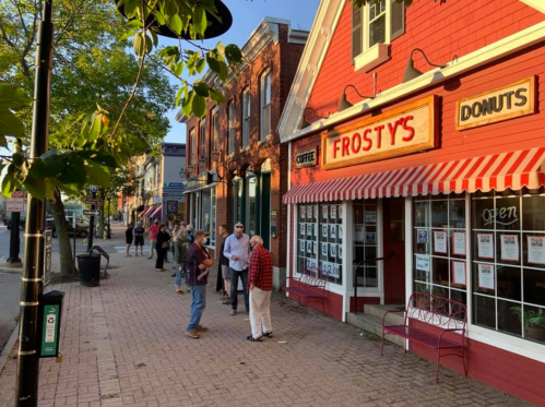 A bustling street scene with people gathered outside Frosty's Donuts, featuring a red building and striped awning.