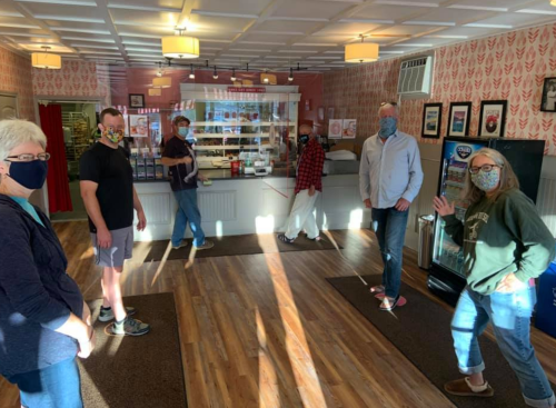 Group of people wearing masks, standing apart in a brightly lit café with a display counter in the background.
