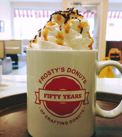 A mug from Frosty's Donuts, topped with whipped cream, caramel drizzle, and chocolate sprinkles, celebrating fifty years.