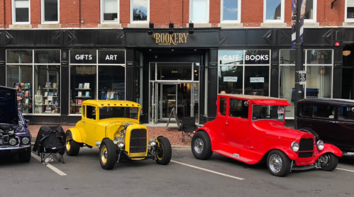 Two vintage cars, a yellow hot rod and a red coupe, parked outside a bookstore with a vibrant storefront.