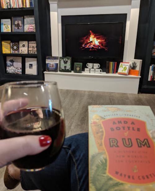 A cozy living room scene with a glass of red wine, a book titled "And a Bottle of Rum," and a fire on the TV.