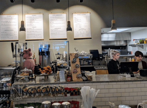 A café interior with a menu board, two employees at the counter, and a display of pastries and drinks.