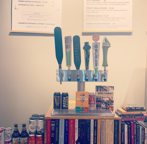 A bar display with beer taps, a menu board above, and a shelf of books and craft beer cans below.