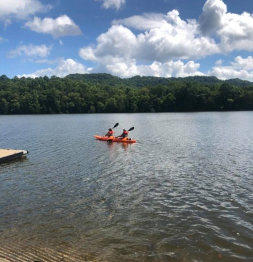 Two people kayaking on a calm lake surrounded by green hills and a blue sky with fluffy clouds.