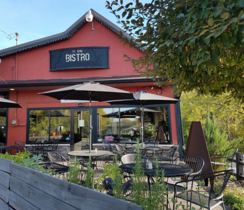 A red bistro building with outdoor seating under umbrellas, surrounded by greenery and trees.