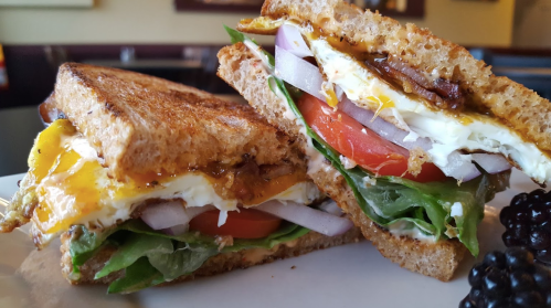 A close-up of a toasted sandwich with layers of egg, tomato, lettuce, onion, and sauce, served with blackberries.