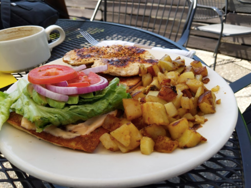 A plate of breakfast featuring a veggie sandwich with lettuce, tomato, and onion, alongside crispy potatoes and coffee.