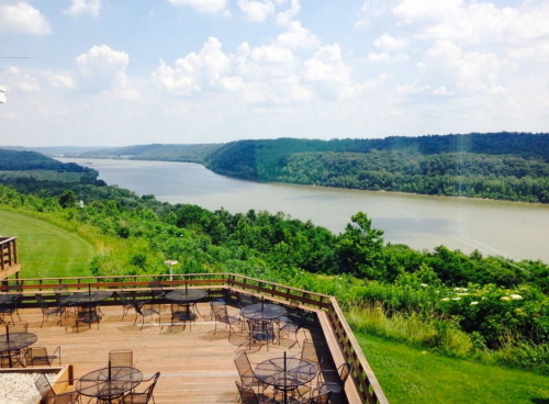 Scenic view of a river surrounded by lush greenery and hills, with a wooden deck and empty tables in the foreground.
