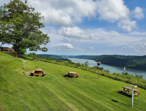 A scenic view of a river surrounded by green hills, with picnic tables on a grassy hillside under a blue sky.