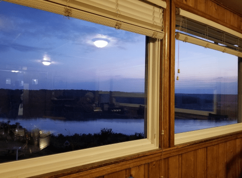 A serene view of a river at dusk, framed by wooden walls and windows, with soft lighting inside.