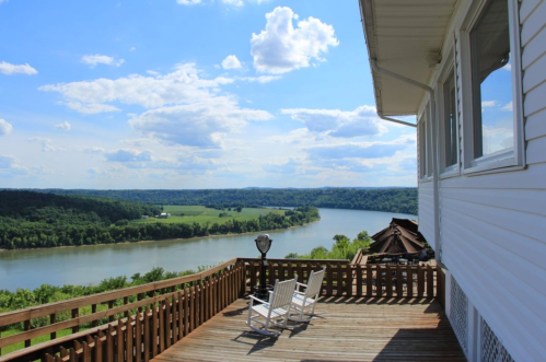 A scenic view of a river from a deck, with white rocking chairs and lush green hills under a blue sky with clouds.