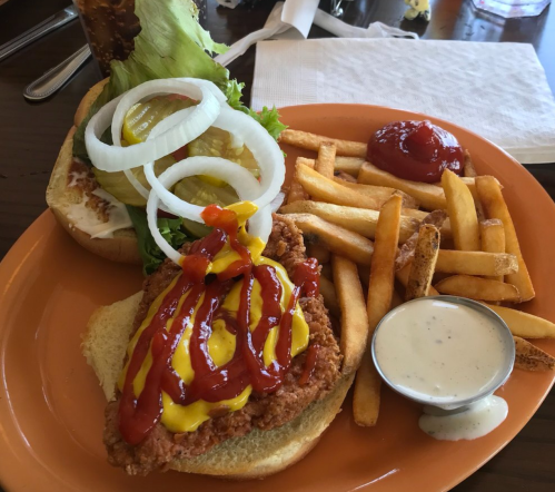A plate with a fried chicken sandwich topped with lettuce, onions, ketchup, and mustard, served with fries and ranch dressing.