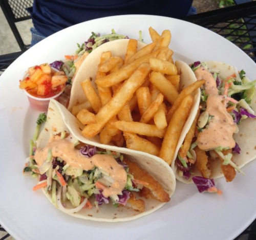 A plate with three tacos filled with slaw and sauce, accompanied by a side of crispy fries and a small fruit cup.
