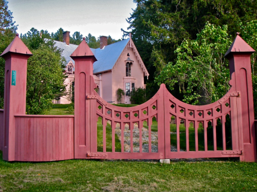 A pink Gothic-style house behind a decorative wooden gate, surrounded by trees and a grassy lawn.