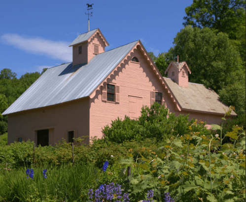 A charming pink barn with a weather vane, surrounded by lush greenery and colorful flowers under a clear blue sky.
