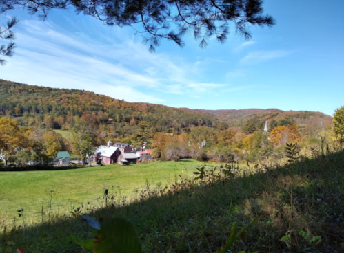 A scenic view of a valley with colorful autumn trees, a farm, and distant hills under a clear blue sky.