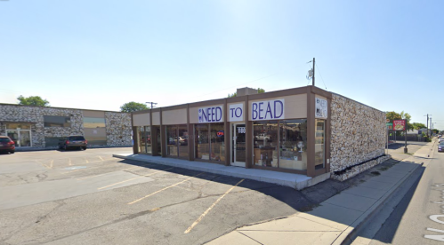 A storefront with a sign reading "Need to Bead," featuring large windows and a stone exterior on a sunny day.