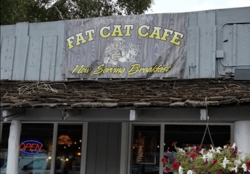 Sign for "Fat Cat Cafe" with a cat graphic, announcing "Now Serving Breakfasts," above a rustic building entrance.