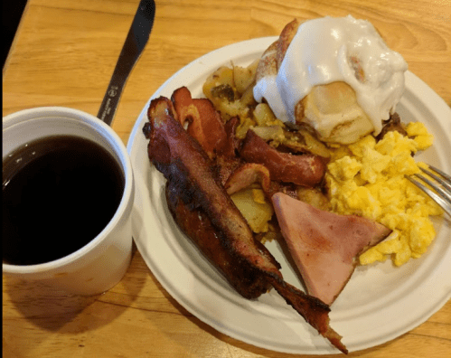 A plate of breakfast featuring scrambled eggs, bacon, ham, potatoes, a cinnamon roll, and a cup of coffee.