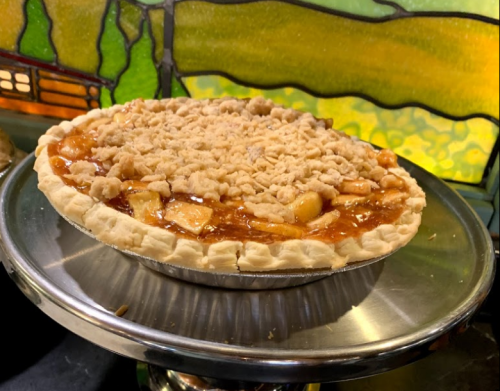 A freshly baked apple pie with a crumbly topping, displayed on a silver cake stand against a colorful stained glass background.