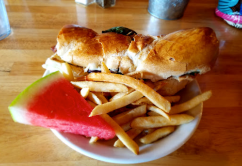 A sandwich with lettuce and tomato, served with a side of French fries and a slice of watermelon on a wooden table.