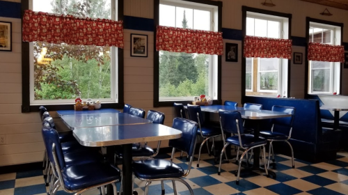 A cozy diner interior with blue chairs, checkered floor, and large windows adorned with red patterned curtains.