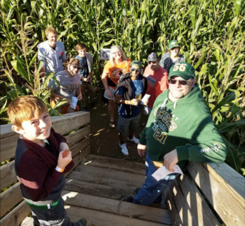 A group of people, including a boy and adults, gather on wooden steps in a corn maze surrounded by tall corn stalks.