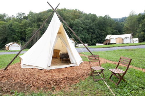 A white tent on a grassy area with wooden chairs nearby, surrounded by trees and additional tents in the background.