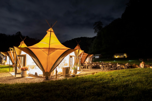 Glowing tents with unique designs set in a grassy area at night, surrounded by trees and outdoor seating.
