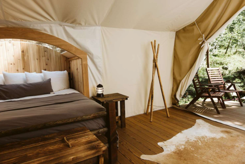 Cozy glamping tent interior with a wooden bed, lantern, and a deck chair on a wooden floor. Natural light filters in.