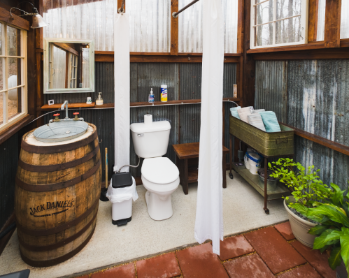 A rustic bathroom with a wooden barrel sink, toilet, and potted plants, featuring corrugated metal walls and natural light.