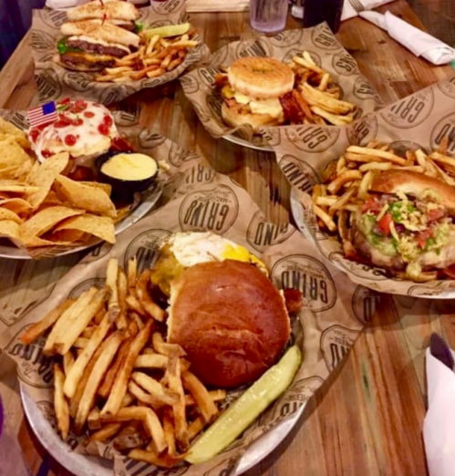A table with five plates of burgers, fries, nachos, and toppings, all served on paper with a rustic wooden background.