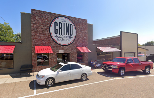 Exterior of The Grind restaurant featuring a brick facade, red awnings, and parked cars in front on a sunny day.