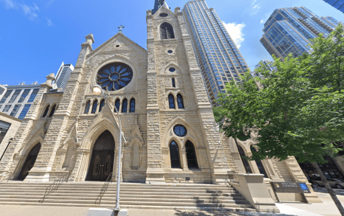 Historic stone church with a large rose window, surrounded by modern skyscrapers and greenery under a clear blue sky.