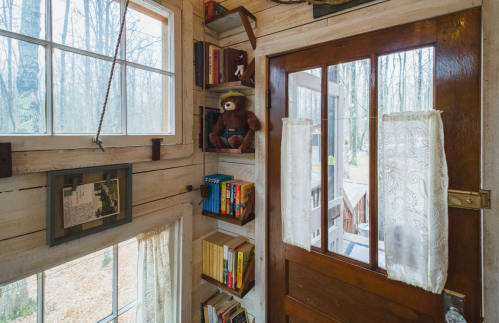 Cozy interior of a cabin with wooden walls, a door, bookshelves, and a teddy bear on display. Natural light filters in.