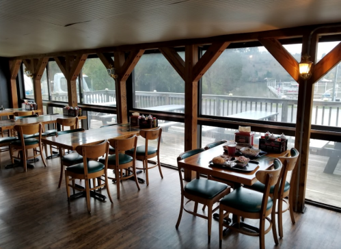 Cozy restaurant interior with wooden beams, tables, and large windows overlooking a marina.